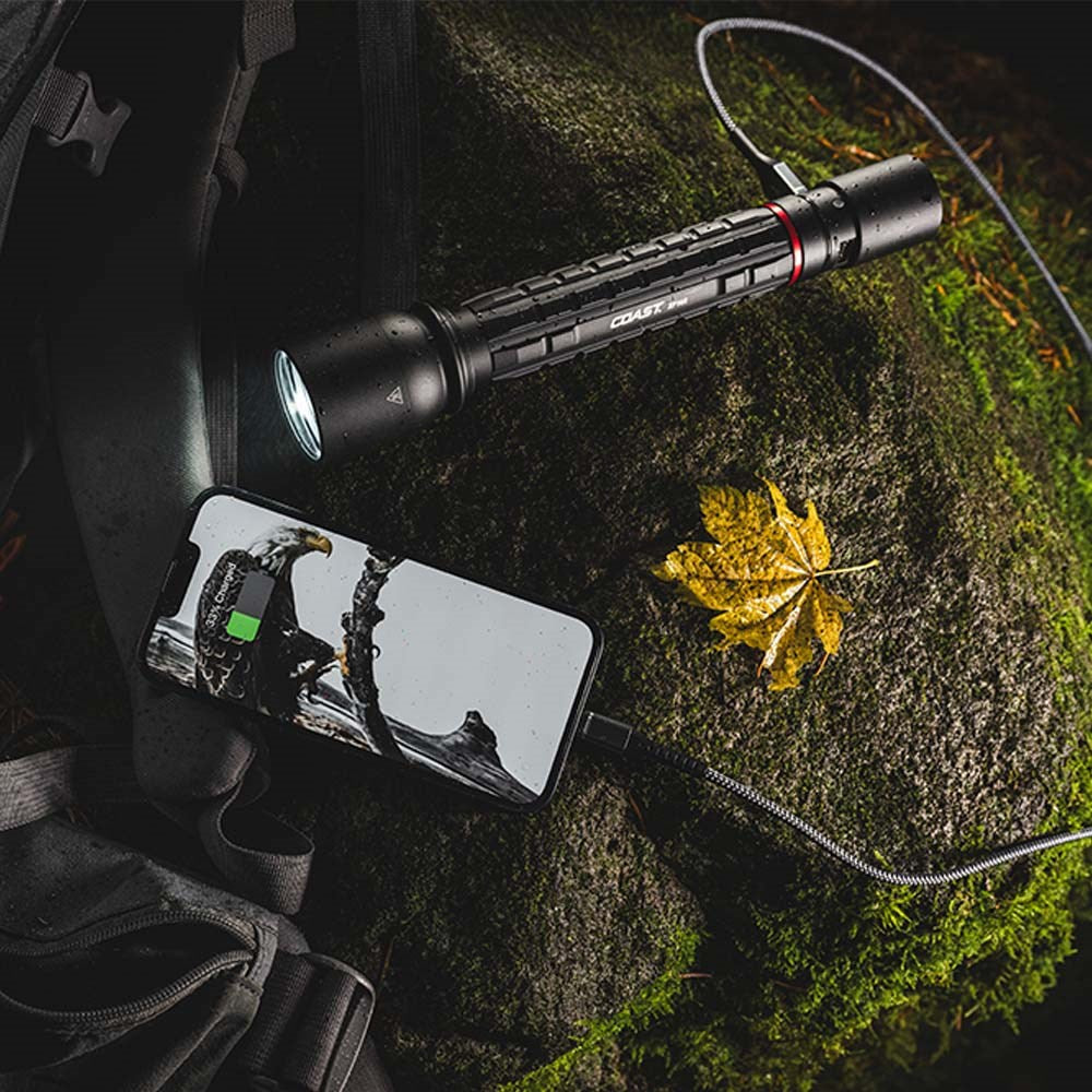 Coast XP14R  Flashlight charging a phone on a mossy surface with a backpack nearby